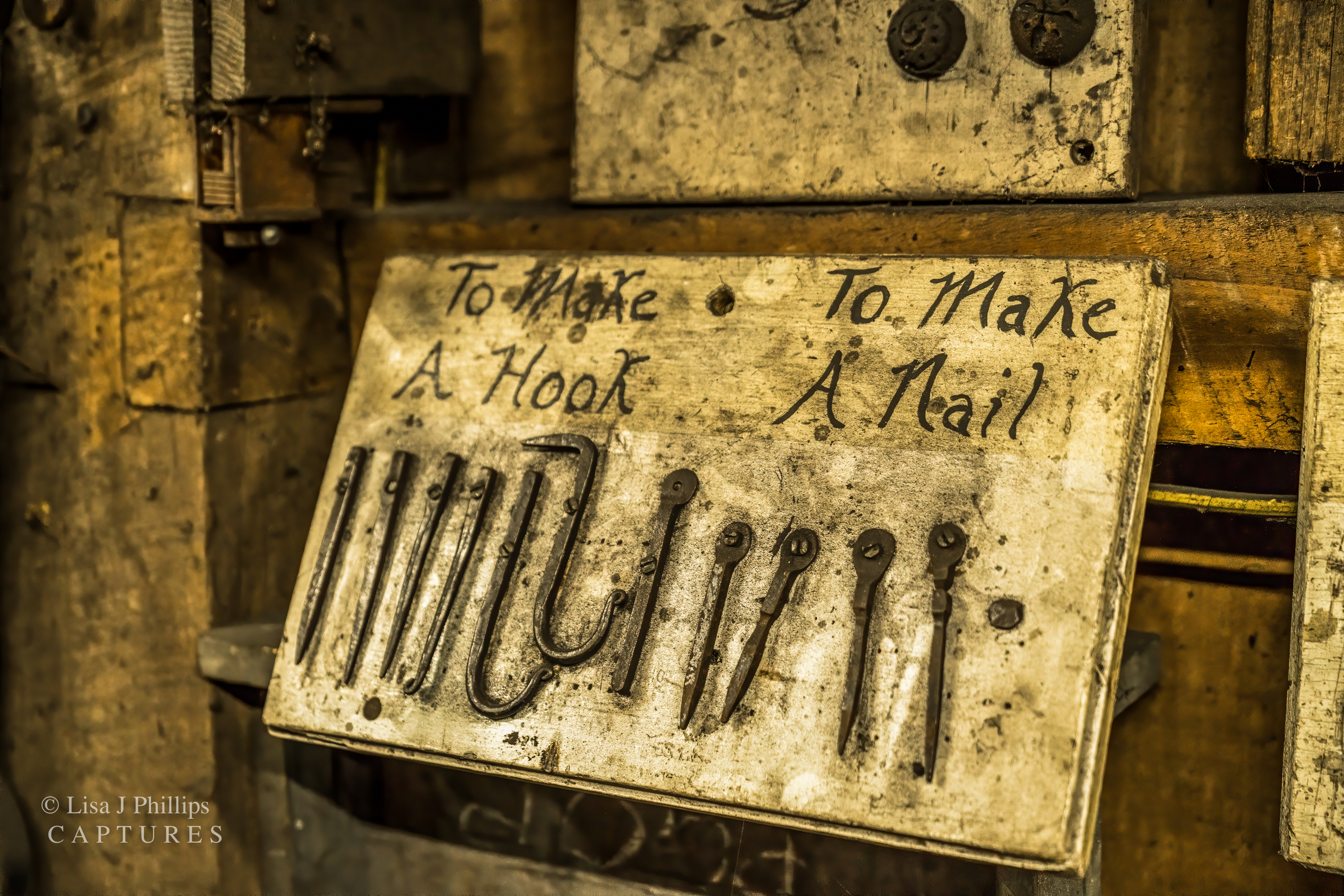 A wooden board leaning against a wall. It reads &lsquo;To Make a Hook&rsquo; on the left side and &lsquo;To Make A Nail&rsquo; on the right. Below are six pieces of steel on the left side showing the steps to make a hook and four pieces of steel on the right side showing the steps to make a nail
